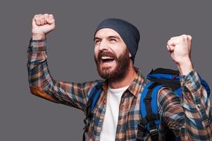 I did it Happy young bearded man carrying backpack and raising his arms up while standing against grey background photo