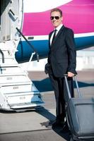 Ready to business trip. Confident mature businessman carrying suitcase and smiling while standing near the airplane entrance photo