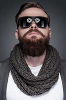 Cool and trendy. Portrait of handsome young bearded man in sunglasses standing against grey background photo