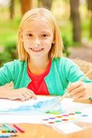 I love drawing Cute little girl drawing something on paper and smiling while sitting at the table and outdoors photo
