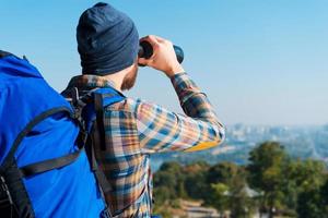 Amazing view. Handsome young bearded man carrying backpack and looking through binoculars at view photo