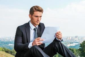 Man examining contract. Confident young man in formalwear examining document while sitting pointing away while standing outdoors with cityscape in the background photo
