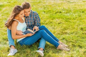 Enjoying their free time together. Beautiful young loving couple sitting on the grass while man pointing book and smiling photo