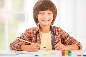 pequeño artista niño feliz relajándose mientras pinta con acuarelas sentado en la mesa foto