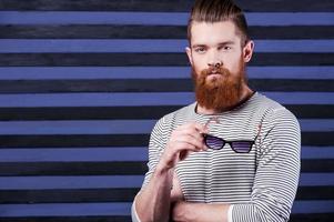 Style in every strip. Handsome young bearded man holding sunglasses in hand while standing against stripped background photo