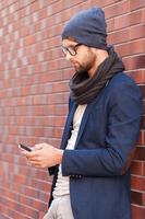 Typing text message. Side view of handsome young man in smart casual wear holding mobile phone while leaning at the brick wall photo