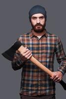 Confident lumberjack. Confident young bearded man holding a big axe and looking at camera while standing against grey background photo