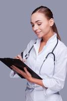 Beautiful doctor at work. Confident female doctor in white uniform writing clipboard while standing against grey background photo