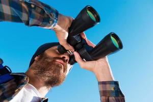 Taking a closer look. Long angle view of handsome young bearded man looking through binoculars away photo