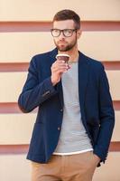 Relaxing with cup of fresh coffee. Handsome young man in smart jacket holding coffee cup and looking away while standing at the street photo