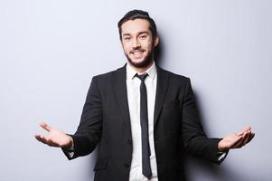 You are welcome Cheerful young man in formalwear looking at camera and gesturing while standing against grey background photo