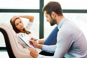 She needs an expert advice. Frustrated young woman sitting at the chair and holding hand on head while young man sitting close to her and writing something in his clipboard photo