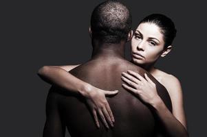 Feeling confident and protected. Beautiful Caucasian woman hugging black man and looking at camera while both standing against grey background photo