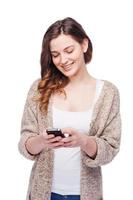 Typing message to friend. Cheerful young woman holding mobile phone and smiling while standing isolated on white photo