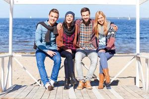 We are always together. Full length of four young happy people bonding to each other and smiling while sitting on the beach photo