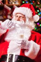 I love milk Cheerful Santa Claus holding glass with milk and adjusting his mustache with Christmas Tree in the background photo