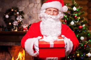 Merry Christmas to you Cheerful Santa Claus stretching out a gift box and smiling with fireplace and Christmas Tree in the background photo