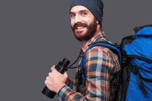 Confident tourist. Side view of handsome young bearded man with backpack holding binoculars and smiling while standing against grey background photo