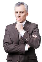 Stern glance of director. Thoughtful mature man in formalwear holding hand on chin and looking at camera while standing against white background photo