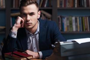 I need inspiration. Thoughtful young author sitting at the typewriter and looking at camera with bookshelf in the background photo