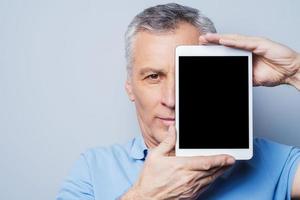 My assistant  in any cases. Confident senior man holding his digital tablet in front of his face and smiling while standing against grey background photo