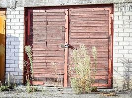 Wide brown wooden door locked with a padlock with grass growing in front of the entrance with midday shadows photo