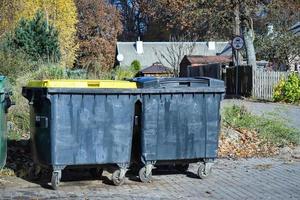 Two black garbage containers on wheels standing on pathway in autumn partk with visible house photo