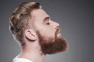 Bearded handsome. Side view of handsome young bearded man keeping eyes closed while standing against grey background photo