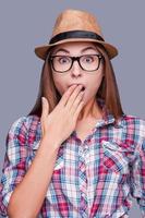 It can not be truth Surprised young woman in glasses and funky hat covering mouth with hand and looking at camera while standing against grey background photo