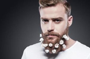 Flowers in my beard. Portrait of handsome young man with flowers in his beard looking at camera while standing against grey background photo