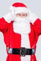 Tired Santa. Frustrated Santa Claus holding head in hands and keeping eyes closed while standing against grey background photo