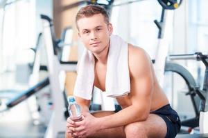 Having a break after workout. Handsome young muscular man holding bottle with water and looking at camera while having a rest in gym photo