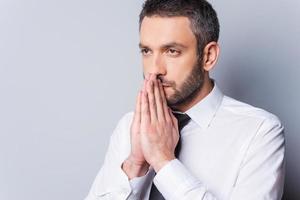 Thinking about solution. Portrait of concentrated mature man in shirt and tie holding hands clasped near face and looking away while standing against grey background photo