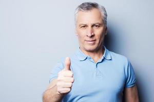 Thumb up for success Cheerful senior man in casual showing his thumb up and smiling while standing against grey background photo