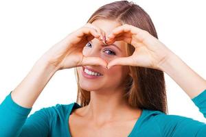 Gesturing finger heart. Beautiful young woman looking at camera and gesturing finger heart while standing isolated on white photo