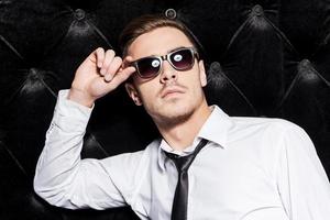 Cool and handsome. Handsome young man in shirt and tie adjusting his sunglasses and looking away while standing against black background photo