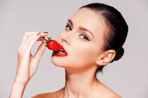 Beautiful young woman holding strawberry in her hand while standing against grey background photo