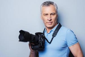 fotógrafo profesional. retrato de un hombre maduro confiado en una camiseta sosteniendo una cámara mientras se enfrenta a un fondo gris foto