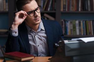 Thoughtful author. Thoughtful young author working at the typewriter and adjusting his eyeglasses while sitting at his working place with bookshelf in the background photo