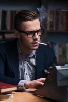 Author at work. Confident young author working at the typewriter and smoking cigarette while sitting at his working place with bookshelf in the background photo