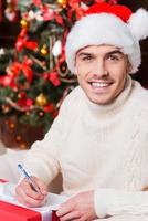 Present for her. Handsome young man in Santa hat writing Christmas letter and smiling with Christmas Tree in the background photo