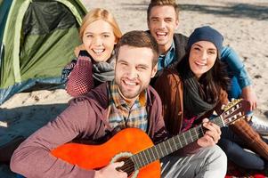 divirtiéndonos juntos. vista superior de cuatro jóvenes felices sentados juntos cerca de la tienda mientras un joven apuesto toca la guitarra y sonríe foto
