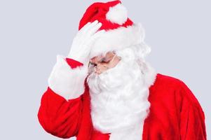 I am so tired. Frustrated Santa Claus holding head in hands and keeping eyes closed while standing against grey background photo