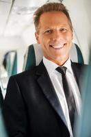sentirse tranquilo y seguro. hombre de negocios maduro confiado sentado en su asiento en el avión y sonriendo foto