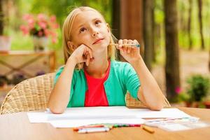 buscando inspiración. niña pensativa sosteniendo la mano en la barbilla y mirando hacia otro lado mientras se sienta en la mesa con lápices de colores y papel sobre ella foto