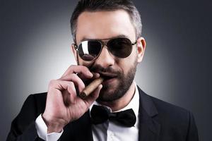 Confident and bossy. Portrait of handsome mature man in formalwear and sunglasses smoking cigar while standing against grey background photo