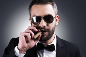 Confident and bossy. Portrait of handsome mature man in formalwear and sunglasses smoking cigar while standing against grey background photo