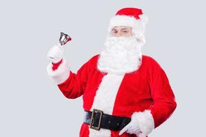 Ho Ho Ho Traditional Santa Claus ringing a bell and looking at camera while standing against grey background photo