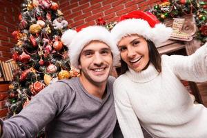 Capturing a happy moment. Beautiful young loving couple bonding to each other and smiling while making selfie with Christmas Tree in the background photo