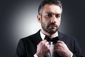 Used to look perfect. Portrait of handsome mature man in formalwear adjusting his bow tie and looking at camera while standing against grey background photo